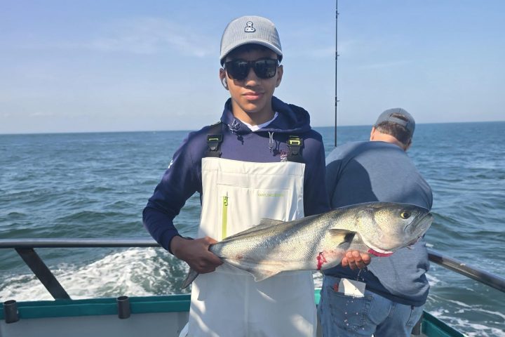 Person in white overalls holding a large fish on a boat, ocean in background.