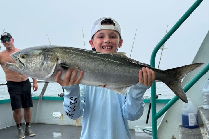 Boy in blue shirt holds large fish on a boat, with a man in the background.