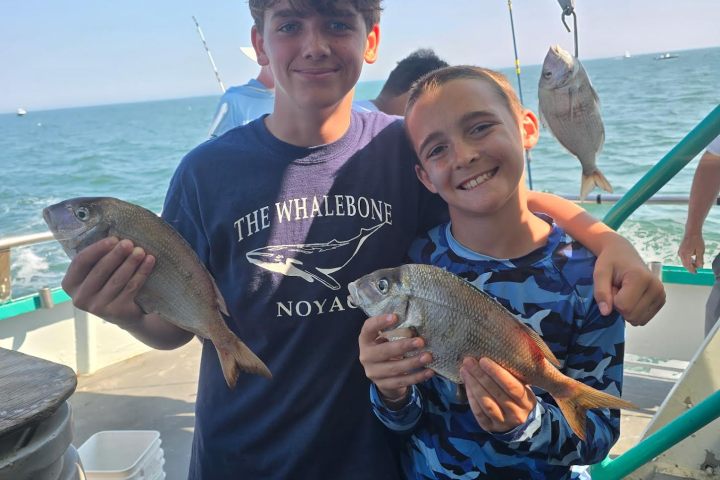Two boys on a boat holding fish, ocean in background.