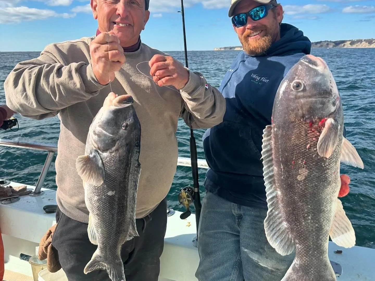 Two men on a boat holding large fish, with the ocean and sky in the background.