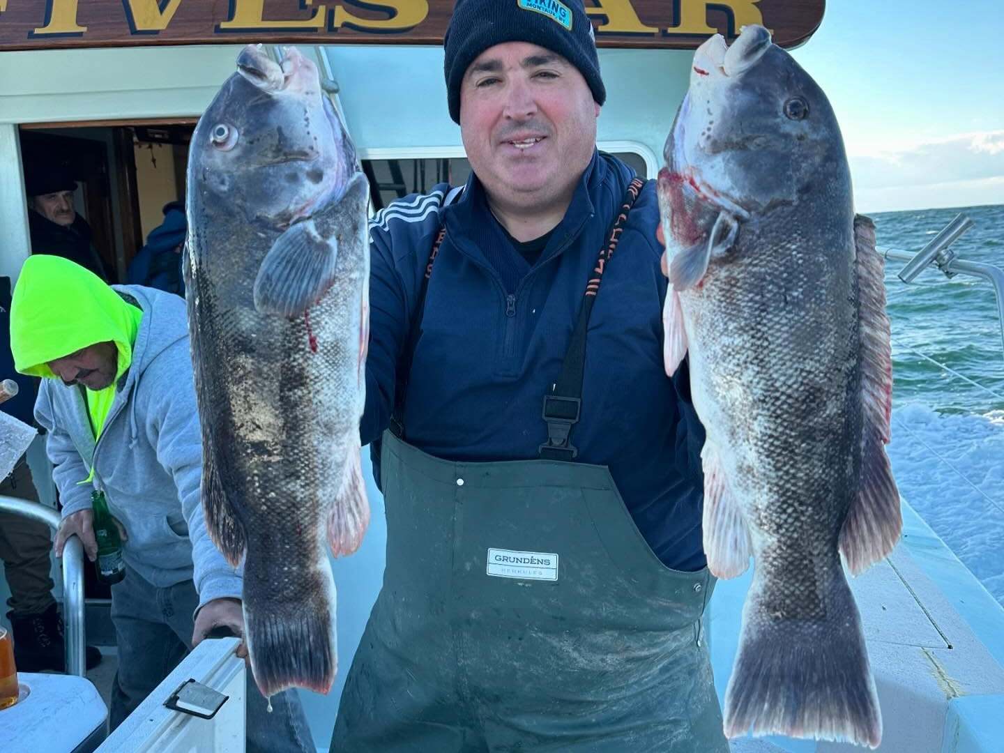 Person on a boat holding two large fish, wearing a beanie and overalls.