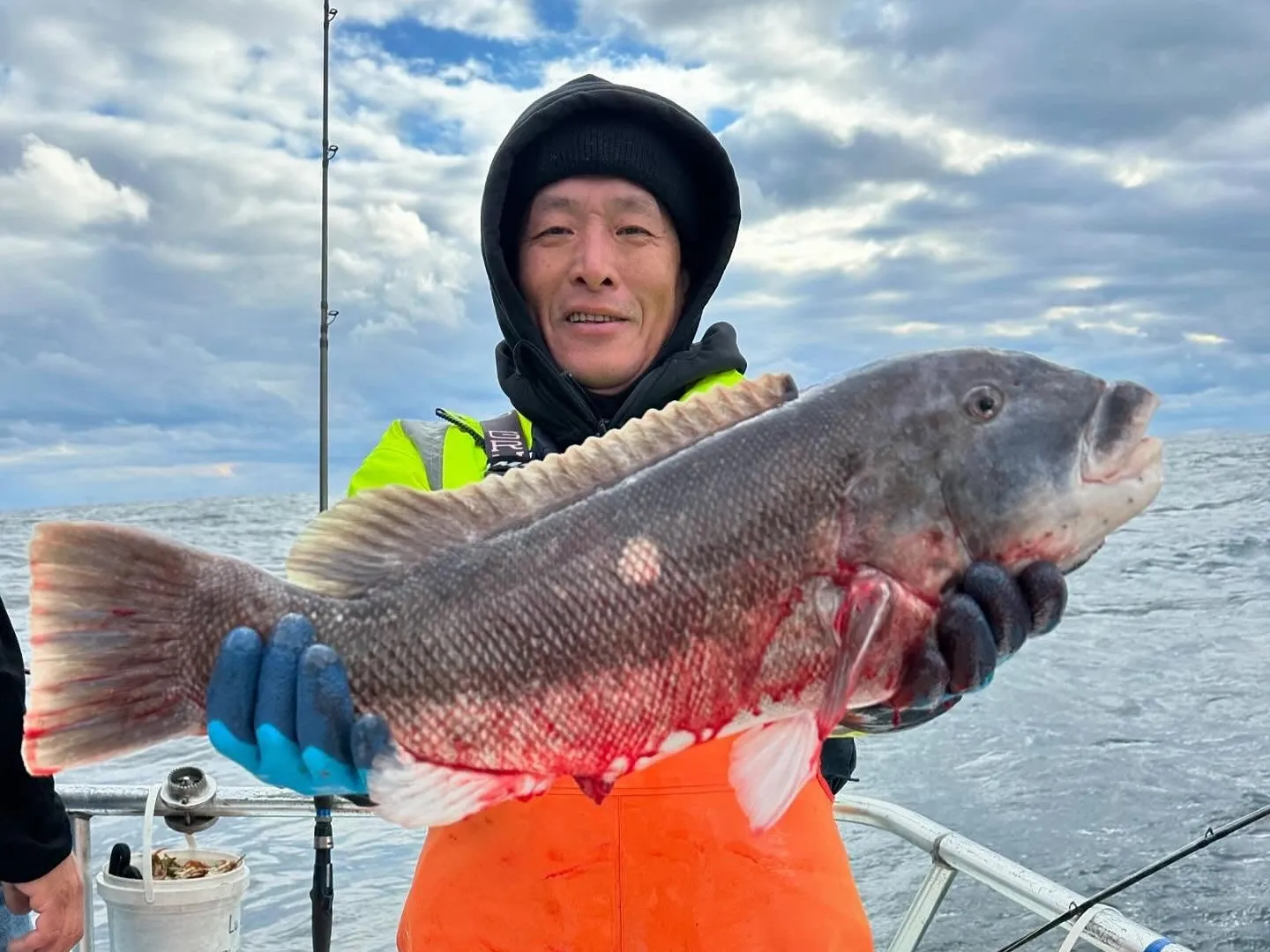 Person on a boat holding a large fish, wearing orange overalls and gloves, with a cloudy sky in the background.
