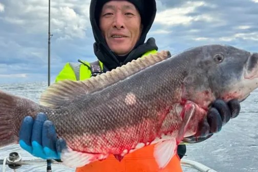 Man in winter gear holds large fish on a boat with cloudy sea backdrop.