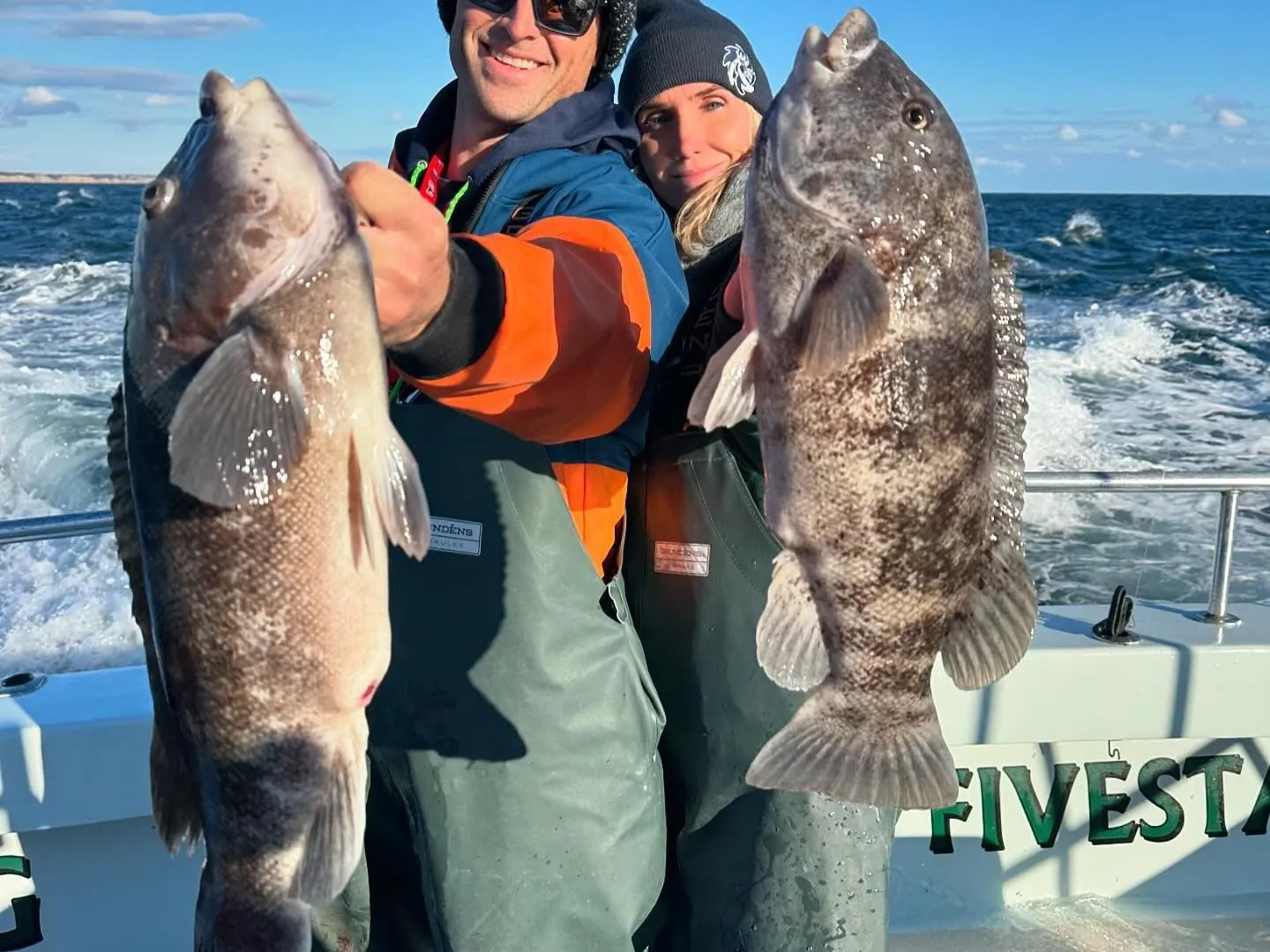 Two people on a boat each holding a large fish, smiling, with the ocean in the background.