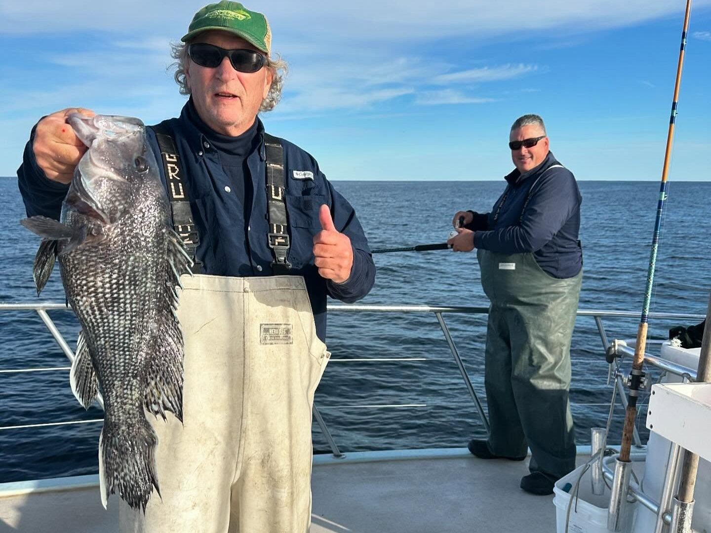 Two men on a boat, one holding a large fish, both wearing fishing gear.
