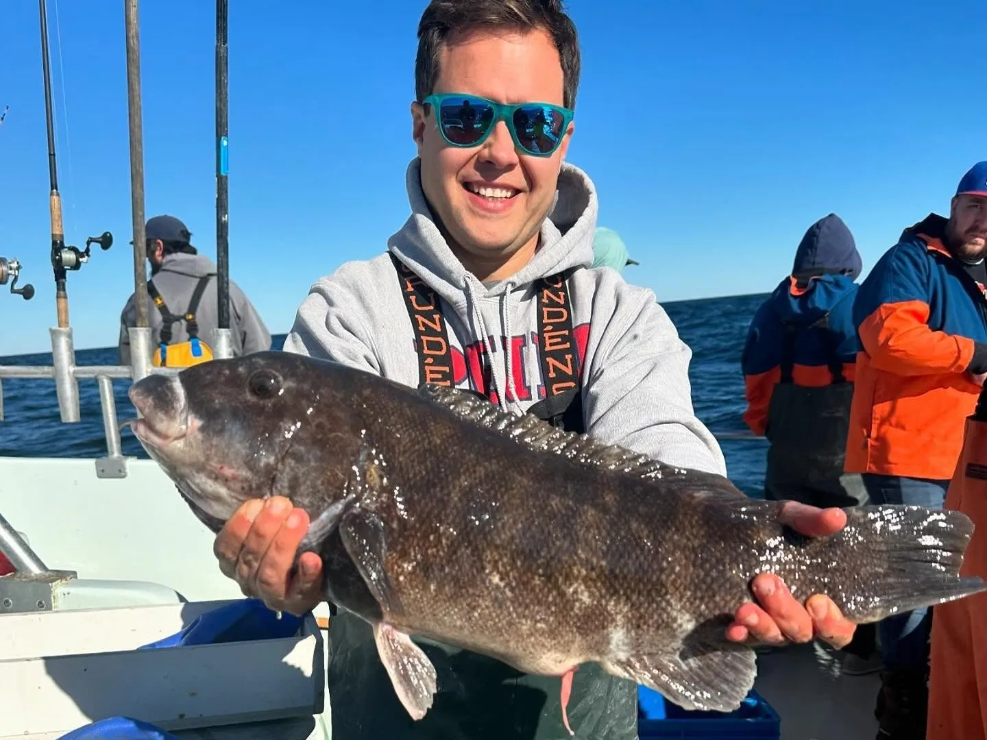 Person on a boat holding a large fish, wearing sunglasses and a hoodie.