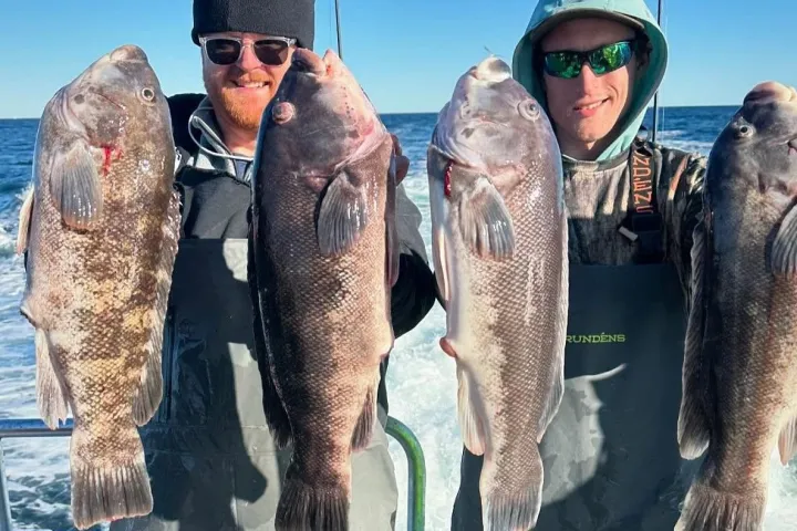 Two people on a boat holding large fish, wearing waterproof gear and sunglasses.