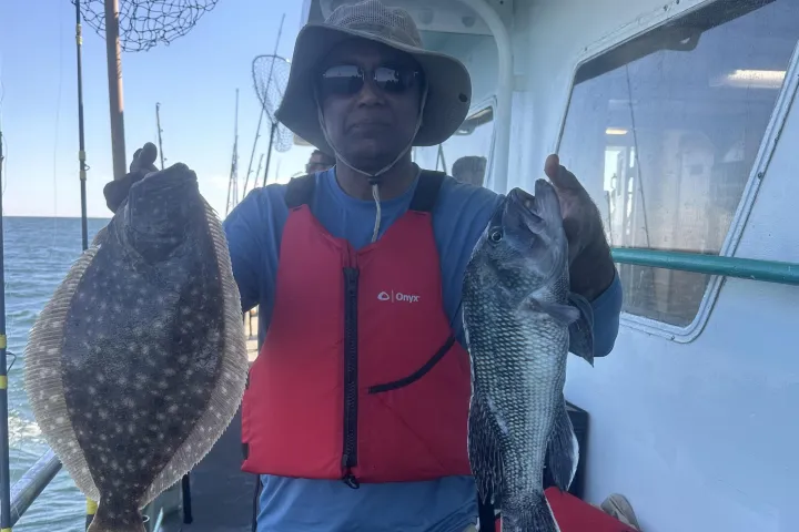 Person on a boat holding a flounder and another fish, wearing a red life jacket and wide-brimmed hat.