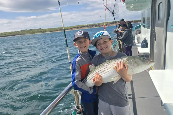 Two boys on a boat holding a large fish with fishing rods and sea in the background.