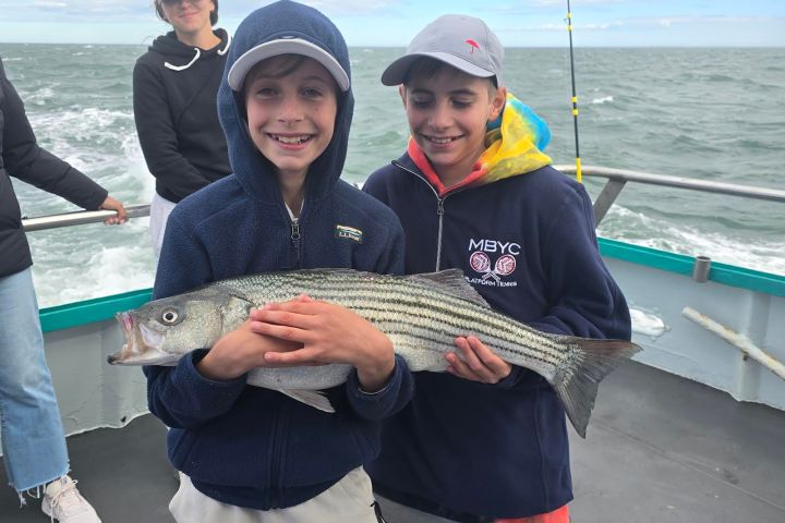 Two kids on a boat holding a large striped fish, smiling, with ocean in the background.