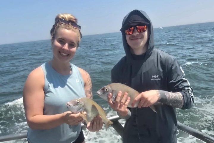 Two people on a boat holding fish with the ocean in the background.