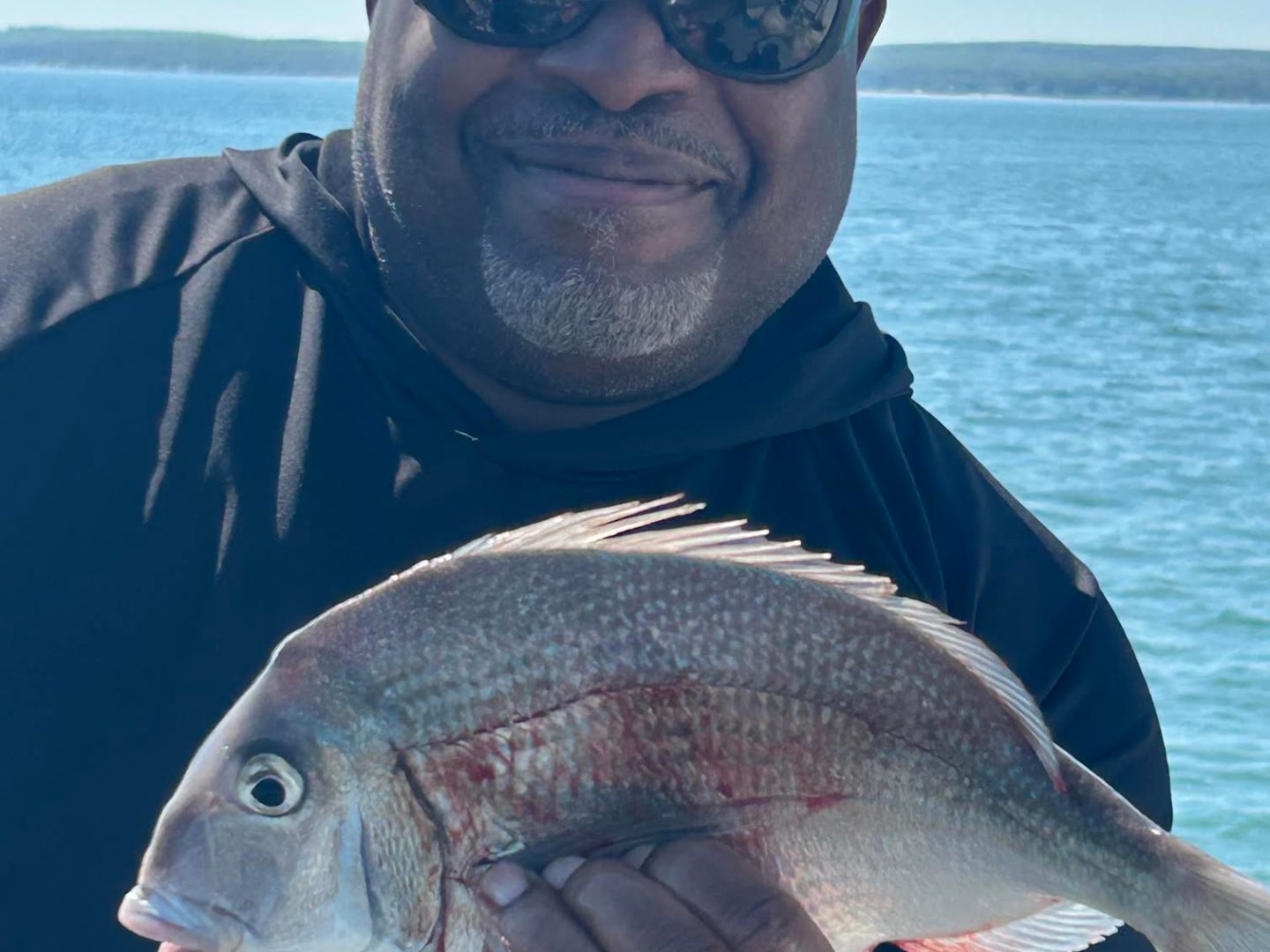 Man holding a fish by the water with sunglasses and a hat.