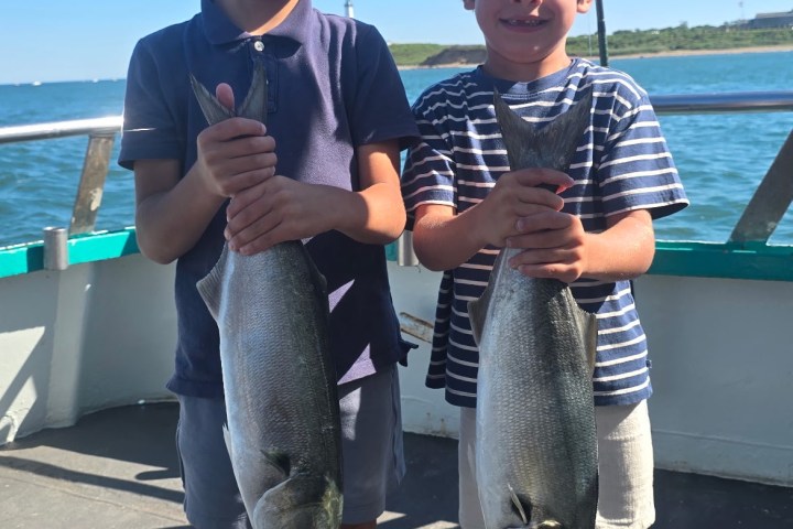 Two children on a boat holding two large fish with blue water in the background.