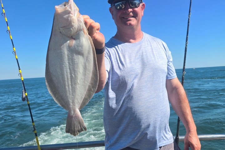 Person holding a large fish on a boat with ocean background and fishing rods visible.
