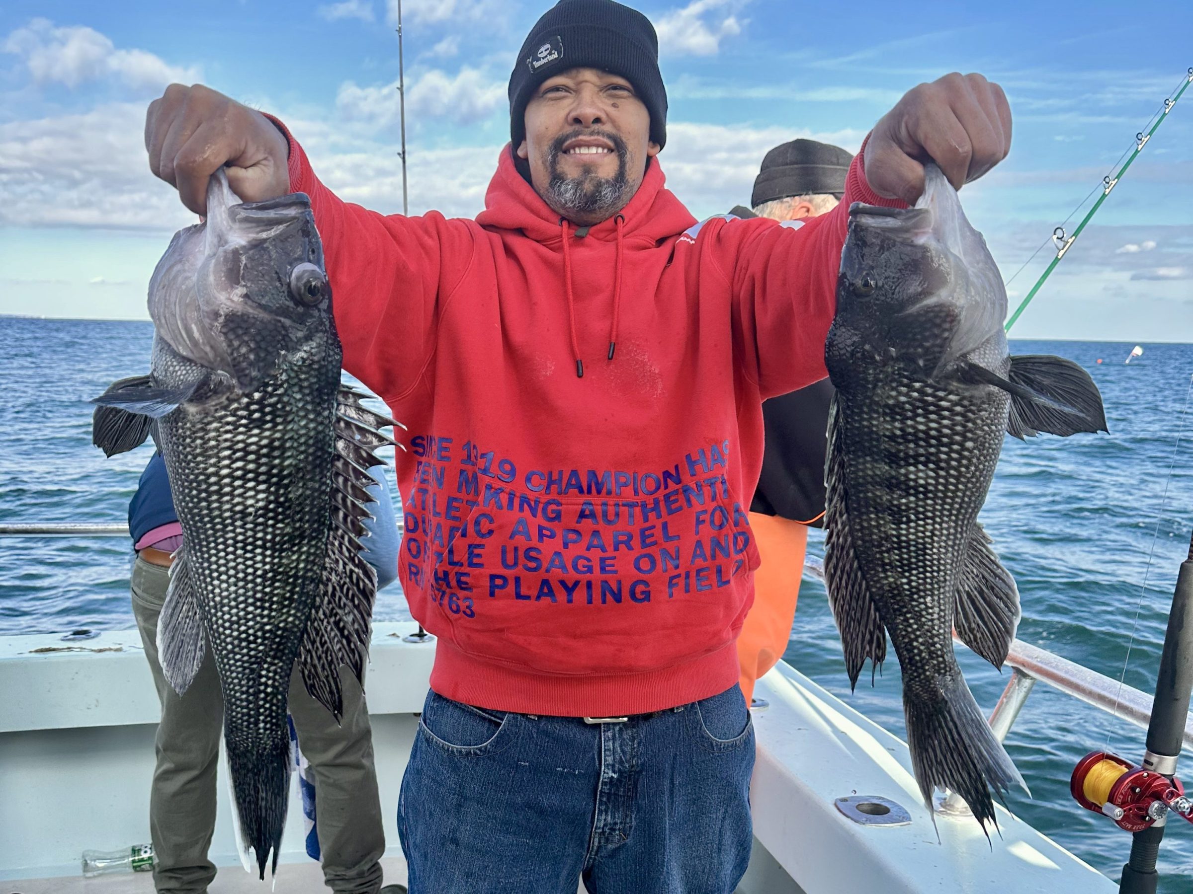 Smiling man on boat holding two large fish, wearing red hoodie and black beanie.