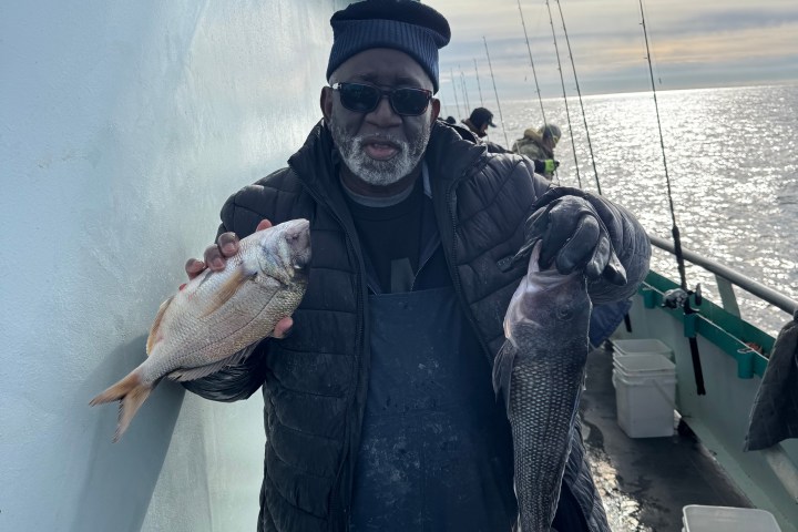 Man holding two fish on a boat with ocean and fishing rods in the background.