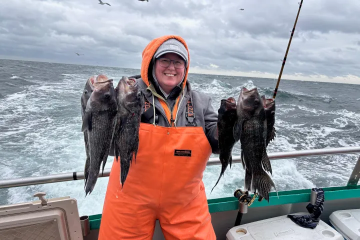 Person on a boat holding multiple fish, wearing an orange suit and smiling, with cloudy sky and sea in background.