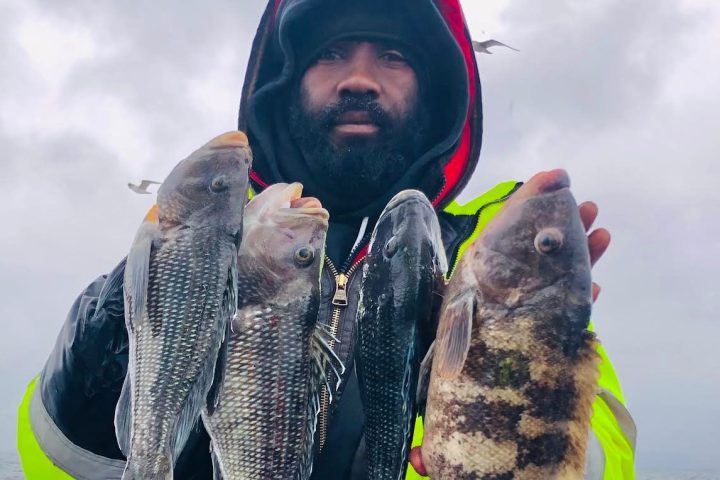 Person in a hooded jacket holding four fish on a boat in cloudy weather.