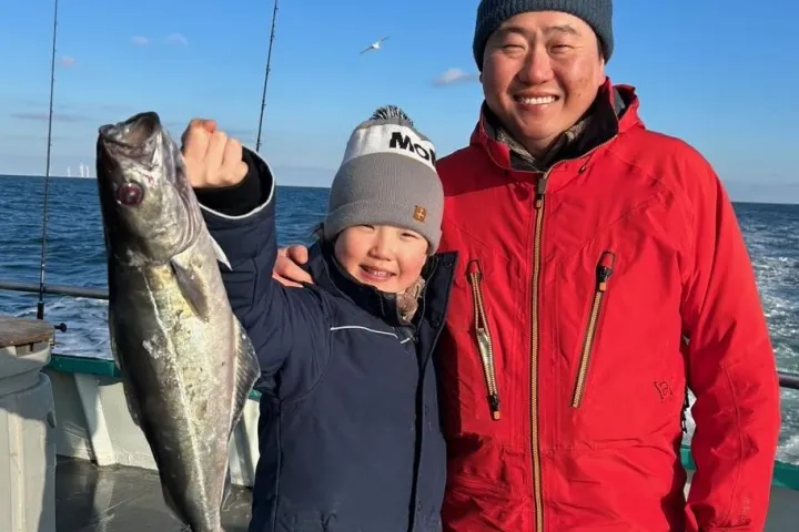 Two people on a boat, one holding a fish, both smiling. Sea and sky with birds in the background.
