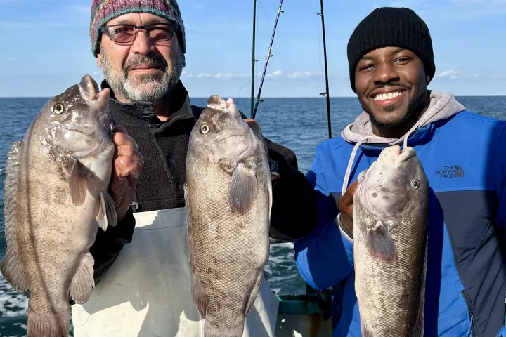 Two men on a boat holding fish, smiling, with sea and sky background.