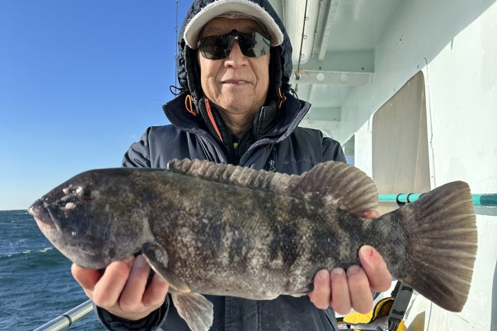 Person on a boat holding a large fish, wearing sunglasses and winter gear, with a clear blue sky.
