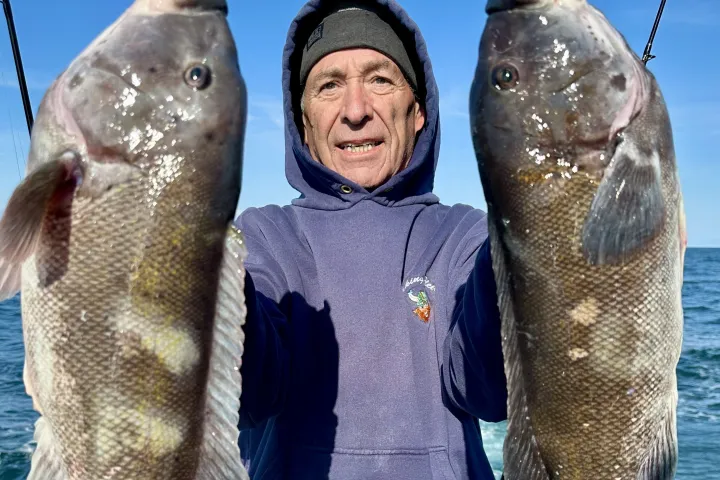 Person in hoodie holds two large fish on a boat with ocean in the background.