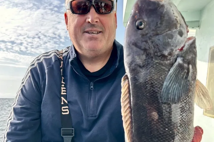 Man in sunglasses holding a large fish on a boat with ocean in the background.