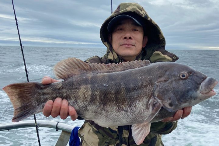 Person in camo jacket holding a large fish on a boat, with ocean and fishing poles in the background.