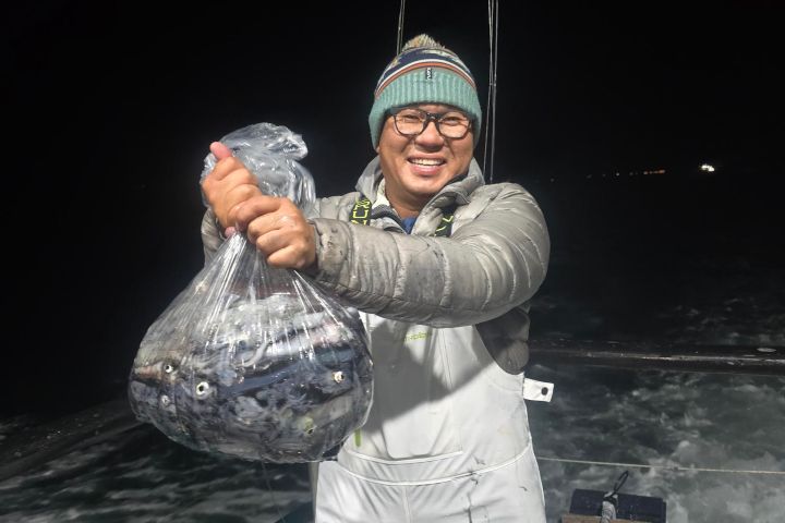 Person on a boat holding a bag of fish at night, wearing a beanie and glasses.