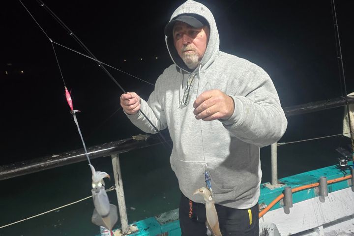 Person in hoodie holds two squids on a fishing line on a boat at night.