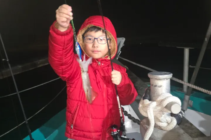 Child in red jacket holds up a small fish on a boat at night.