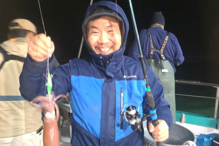 Man holding a fishing rod and a squid on a boat at night, wearing a blue jacket.