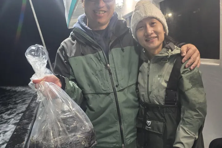 Two people in fishing gear on a boat at night, one holding a bag.