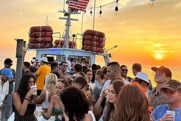 People partying on a boat at sunset, with string lights and an American flag.