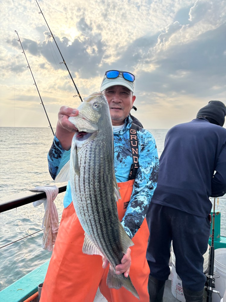 Person holds a large fish on a boat with fishing rods and cloudy sky.