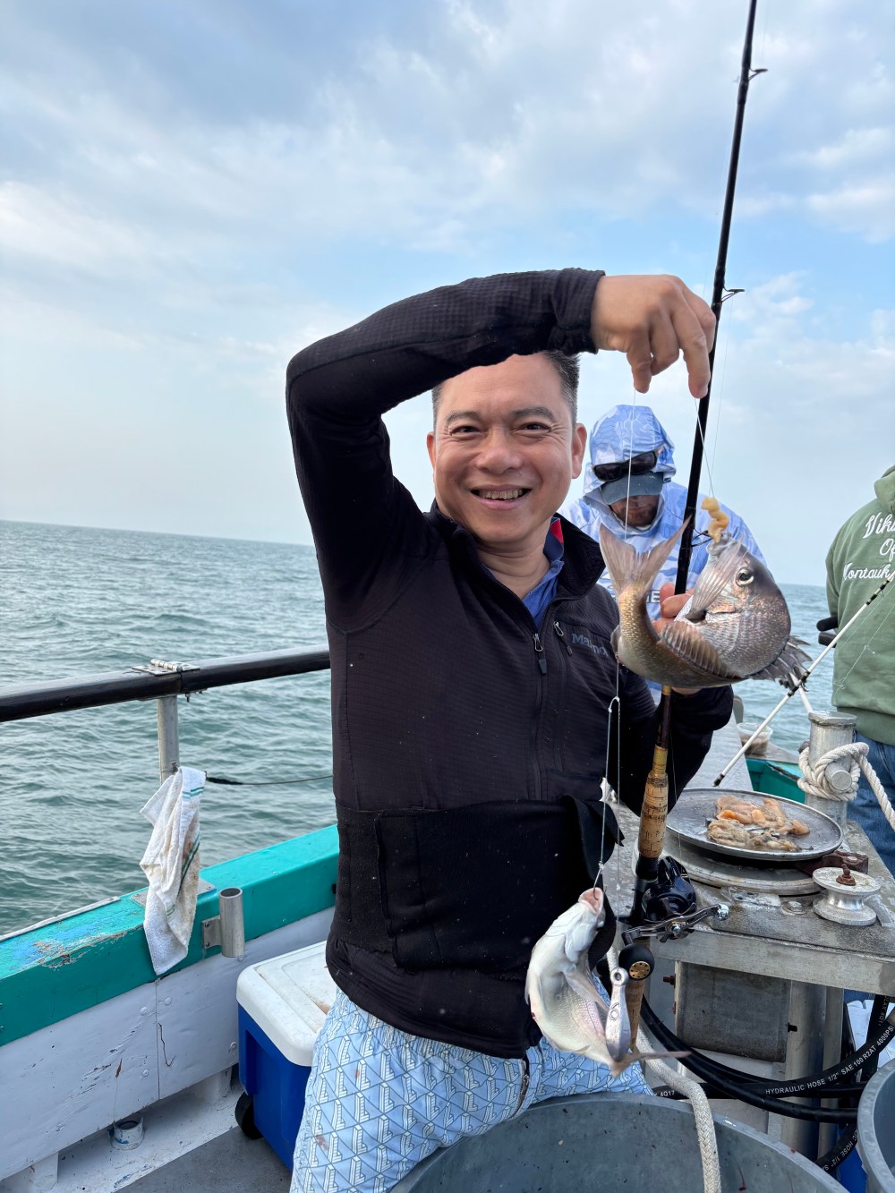 Man smiling, holding fishing rod with two caught fish on a boat.